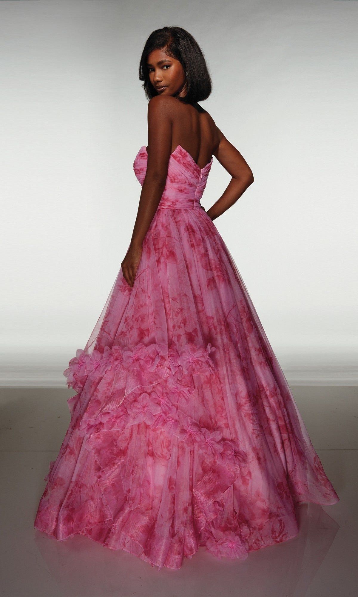 A woman poses with her back partially turned, wearing the Alyce 62042 long prom dress—a strapless pink floral ballgown featuring a ruffle skirt and 3D flower details—against a plain background.